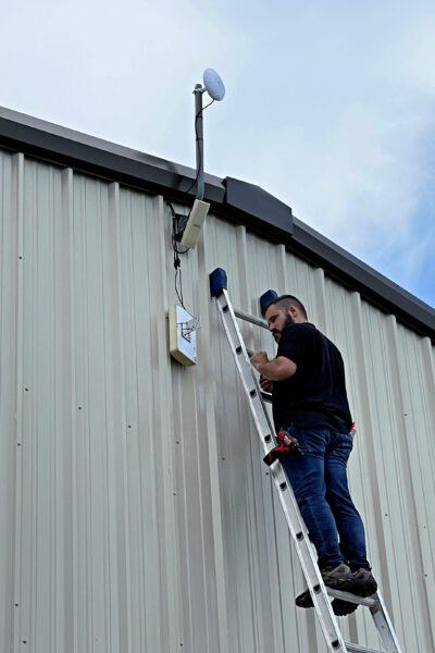 Conlyn standing on a ladder beside a metal building exterior, working on mounted networking equipment, including a directional antenna and associated cabling.