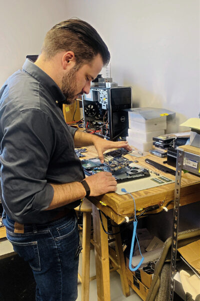 Conlyn standing at a workbench assembling or repairing computer hardware. He is focused on an open motherboard and various components laid out on the bench.
